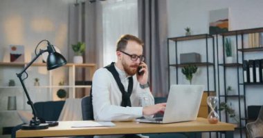 İyi donanımlı bir ofiste bilgisayar başında çalışırken, gözlüklü, telefonla konuşan sevimli, tecrübeli, sakallı bir ofis çalışanı.
