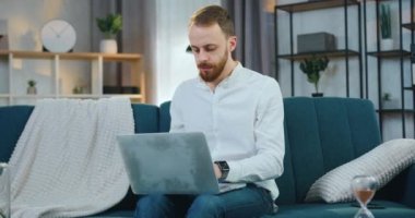 Sevimli, ciddi, 30 yaşında, sakallı, modern bir adam evdeki rahat koltukta oturuyor ve bilgisayardaki farklı raporlar ve verilerle çalışıyor, uzaktan çalışma kavramı.