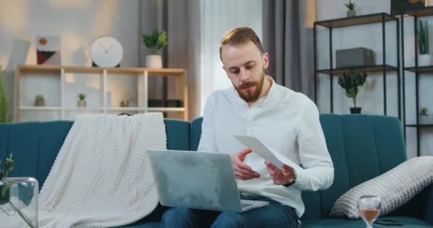 Atraktif terkonsentrasi muda berjenggot pria dalam pakaian santai duduk di sofa lembut di rumah memeriksa laporan dan memasukkan datas ke laptop — Stok Video