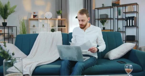 Attraktive selbstbewusste, erfahrene, bärtige Büroangestellte, die zu Hause auf der Couch sitzt und mit Geschäftsdokumenten und Computer arbeitet, hautnah — Stockvideo