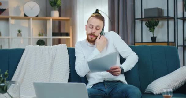 Όμορφος συμπυκνωμένος επιτυχημένος νεαρός γενειοφόρος εργαζόμενος που κάθεται στον καναπέ στο σπίτι και αναθεωρεί τα οικονομικά έγγραφα ενώ μιλάει στο τηλέφωνο με συνάδελφο ή συνεργάτη — Αρχείο Βίντεο