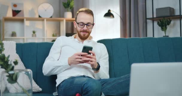 Agradable sonriente joven barbudo feliz hombre en gafas sentado en pose relajada en sofá suave en casa y aplicaciones de teléfono de navegación, concepto de ocio — Vídeos de Stock