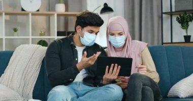 Maskeli yakışıklı, bronzlaşmış bir adam ve koruyucu maskeli Arap kız arkadaşı, evde oturup Cavid-19 hakkında Tablet 'te okudukları bilgiler hakkında konuşuyorlar.