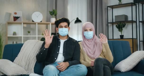 Привлекательная молодая арабская пара в медицинских масках сидит дома на мягком диване во время изоляции коронавирусной пандемии и машет руками в камеру — стоковое видео