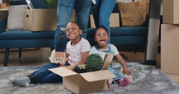 Schöne glücklich lächelnde kleine zwei afrikanisch-amerikanische Mädchen sitzen auf dem Boden mit Karton voller unterschiedlicher Dinge in der Nähe ihrer Eltern am Umzugstag in neue Wohnung — Stockvideo