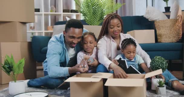 Concepto de bienes raíces donde atractiva sonriente familia afroamericana feliz con los niños que se mudan a un nuevo apartamento y desembalaje caja de cartón wiith decoración del hogar — Vídeos de Stock