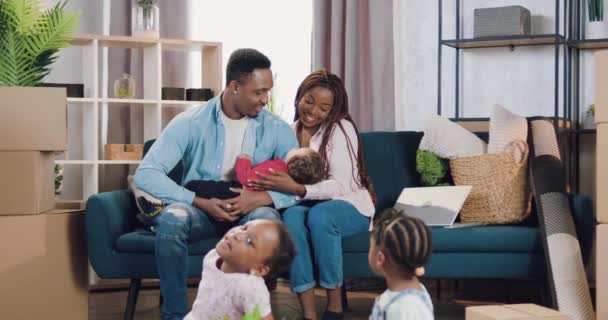 Schön glücklich lächelnde junge afrikanisch-amerikanische Familie mit kleinen Kindern zog in das neu erworbene Haus und sitzt mit dem kleinsten Baby auf dem Sofa, während zwei weitere Kinder auf dem Boden spielen — Stockvideo
