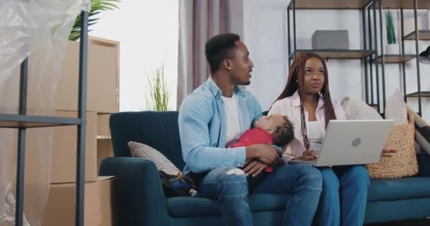 Όμορφος χαρούμενος χαμογελαστός 30χρονος αφροαμερικανός ζευγάρι με το μωρό κάθεται στον καναπέ ανάμεσα σε συσκευασίες από χαρτόνι και χρησιμοποιώντας το laptop σχεδιασμό για την παροχή νεοαποκτηθείσα σπίτι — Αρχείο Βίντεο