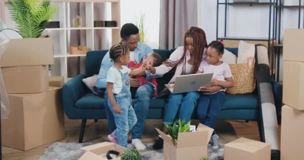 Υπέροχη χαρούμενη, χαρούμενη, νεαρή αφροαμερικάνικη οικογένεια με τρία μικρά παιδιά μετακόμισε σε νέα κατοικία και κάθεται όλοι μαζί στον καναπέ ανάμεσα σε χαρτοκιβώτια. — Αρχείο Βίντεο