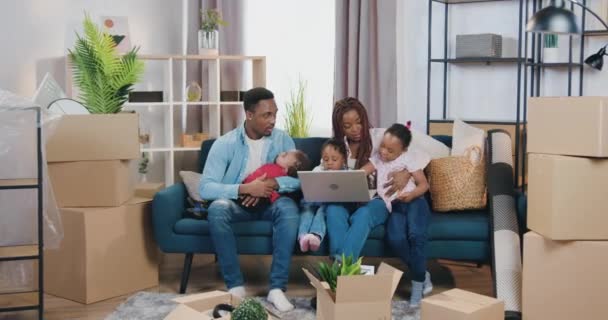 Jolie mère souriante à la peau noire avec dreadlocks assis sur le canapé avec ses deux petites filles et utilisant un ordinateur portable tandis que son mari bercer leur le plus petit fils dans un nouvel appartement — Video