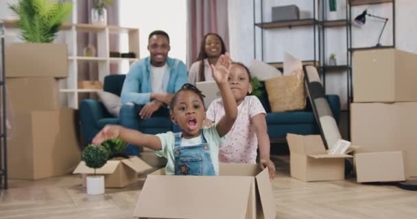 Adoráveis felizes alegres modernos pais de pele preta sentados juntos no sofá em habitação recém-adquirida e assistindo como jogar suas duas filhas em caixa de papelão — Vídeo de Stock