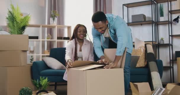 Attrayant joyeux amical aimant couple afro-américain s'amuser avec leurs deux petites filles mignonnes qui se cachent dans l'emballage en carton pendant la journée de déménagement dans une nouvelle maison — Video