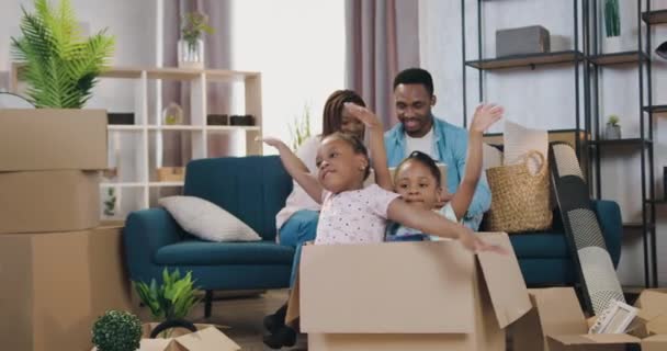 Heerlijk vrolijk lachend modern Afrikaans Amerikaans echtpaar zittend op de bank tussen kartonnen dozen op bewegende dag terwijl hun twee dochtertjes in een kartonnen doos zitten en de vleugels van een vliegtuig afbeelden — Stockvideo