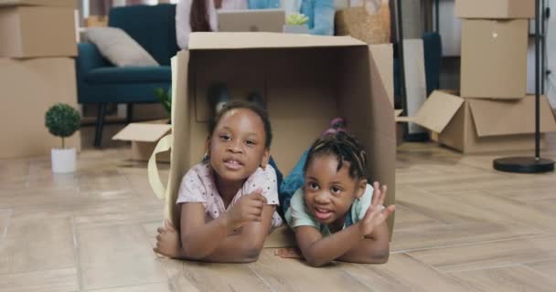 Красивые веселые маленькие две темнокожие девочки играют внутри коробки и машут руками, чтобы поздороваться, глядя в камеру, крупным планом — стоковое видео