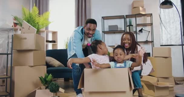 Bom-olhando feliz alegre casal americano africano brincando com suas duas filhas engraçadas que escalando dentro na caixa de papelão depois de desempacotar esta caixa na própria nova habitação — Vídeo de Stock