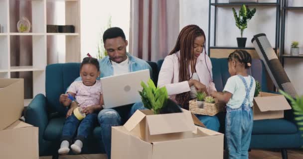 Goed uitziende glimlachende, moderne, jonge, donkere familie met kleine kinderen die hun vrije tijd doorbrengen in nieuw verworven huis, vader met een dochter die iets op de laptop bekijkt, moeder met een andere. — Stockvideo