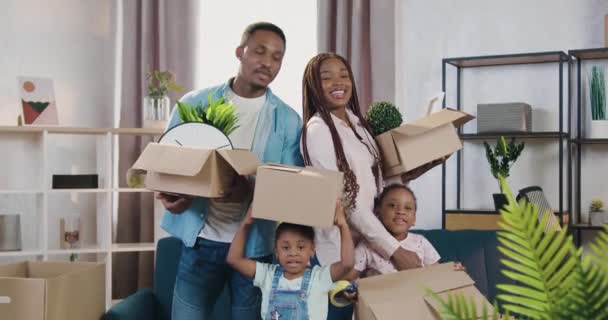 Jóképű, vidám, modern, fekete bőrű család kisgyerekekkel, akik dobozos dobozokat tartanak, lakberendezéssel és elégedett arcú kamerába néznek a saját új lakásukban. — Stock videók