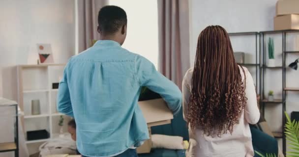 Vista trasera de la encantadora y sonriente pareja afroamericana joven satisfecha que lleva cajas de cartón en su nuevo apartamento después de la reubicación — Vídeos de Stock