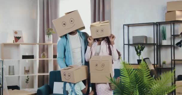 Casal afro-americano despreocupado moderno engraçado irreconhecível e seus filhos se divertindo juntos em nova habitação própria ao colocar em caixas de papelão em suas cabeças em que sorrisos retratados — Vídeo de Stock