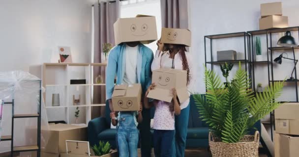 Zabawnie wyglądająca nowoczesna rodzina z dwójką małych dzieci chowających głowy pod kartonowe pudełka z przedstawionymi uśmiechami wśród innych pudełek po przeprowadzce do nowego mieszkania — Wideo stockowe