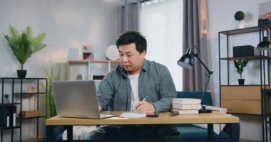 İyi görünümlü, çalışkan, Asyalı bir yetişkin adam masada oturuyor ve bilgisayarda çalışıyor. Evinde, ön tarafta özel olarak tasarlanmış bir yerde.