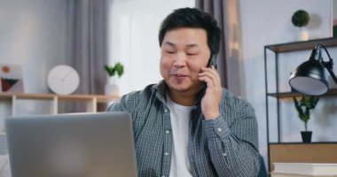 Güzel görünüşlü gülümseyen modern Çinli adamın portresi. Ev bilgisayarının önünde oturuyor ve cep telefonuyla konuşuyor.
