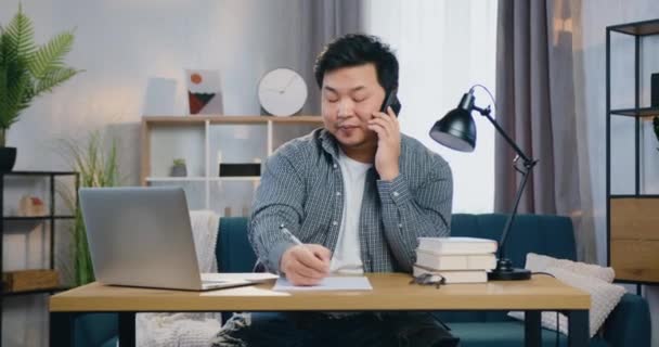 Guapo positivo éxito adulto moderno asiático hombre sentado en su lugar de trabajo en casa y tiene teléfono conversación simultáneamente escribir notas en papel informe — Vídeo de stock