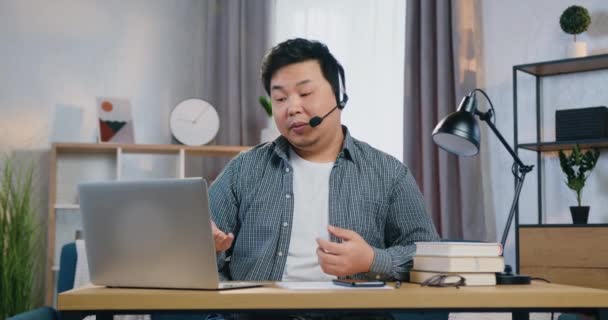 İyi görünümlü, kendine güvenen, kulaklıklı genç Asyalı adam. İşvereniyle video görüşmesi sırasında evde dizüstü bilgisayarın önünde oturuyor. Covid-19 pandemisi açısından uzaktan görüşme. — Stok video
