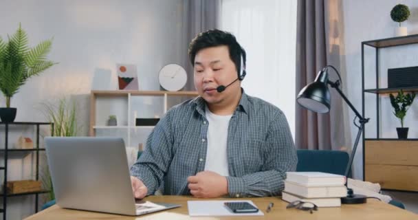 Probable sourire confiant jeune homme chinois en casque assis à la table dans le bureau à la maison et regardant la caméra pendant la vidéoconférence avec des collègues ou des clients, concept de travail à distance — Video