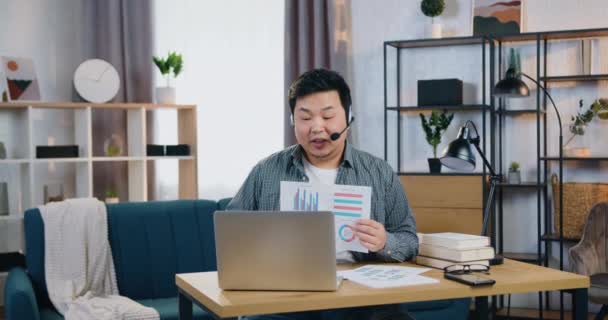 Fešák zkušený sebevědomý mladý asijský muž ve sluchátkách sedí v domácí kanceláři v blízkosti notebooku a pořádá on-line setkání vysvětlující výsledky vyrobené práce pomocí diagramů — Stock video