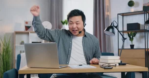 Snygg glad nöjd 30-årig kinesisk kontorsarbetare i headset sitter framför laptop och överraskad av goda nyheter på skärmen och höja handen — Stockvideo