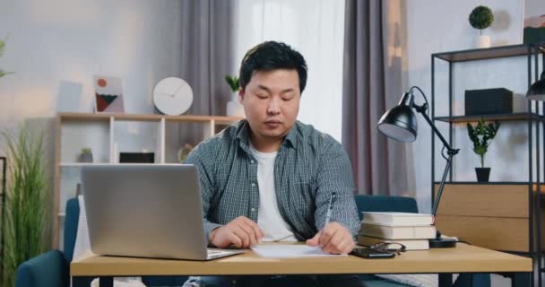 Привлекательный опытный азиат, сидящий дома на работе и работающий над ноутбуком, выписывающий необходимые данные на бумаге для своего бизнес-проекта — стоковое видео