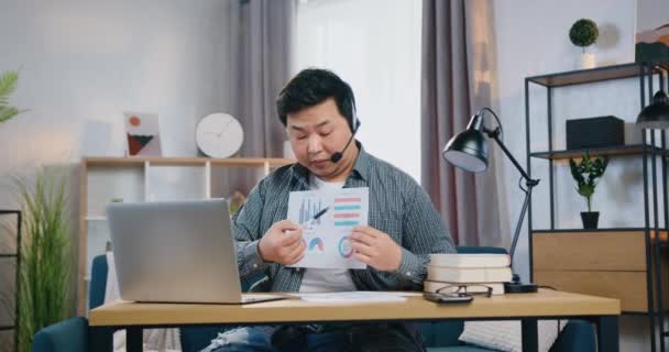 Vonzó céltudatos tapasztalt fiatal ázsiai férfi fülhallgatóban ül a laptop előtt, és elmagyarázza a diagramokat partnereinek az online videokonferencia során, távmunka koncepció — Stock videók