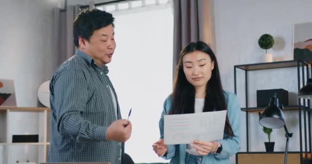 Knappe glimlach tevreden slimme 30-jarige aziatische man uit te leggen resultaten van gemaakt werk met behulp van grafieken aan zijn jongere vrouwelijke collega tijdens gezamenlijke werk over project — Stockvideo