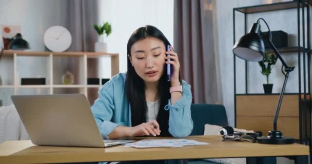 Retrato de encantadora chica asiática inteligente positiva moderna que habla por teléfono mientras trabaja en el ordenador portátil y diferentes informes, cámara lenta — Vídeo de stock