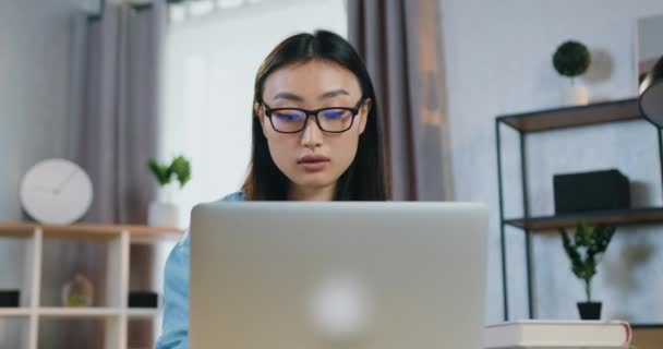 Güzel, konsantre olmuş, gözlüklü Asyalı genç kız ev ofisinde dizüstü bilgisayarda çalışıyor. — Stok video