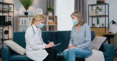 Sağlık ve tıp konsepti. Çekici, sarışın, maske takan kadın doktor hastasını evinde ziyaret ediyor ve hastalık belirtilerini dinliyor.