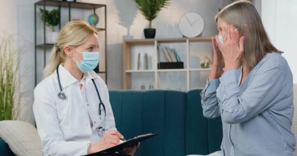 Mulher doente de meia-idade bonita em máscara facial conversando com médico local fêmea em máscara protetora também que visitá-la em casa durante a doença — Vídeo de Stock
