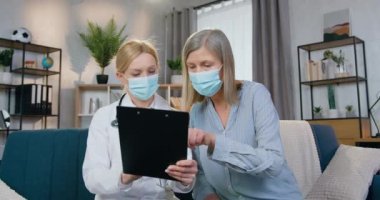 Üzgün, 50 yaşında, tıbbi maskeli bir kadın yerel sağlık görevlisiyle birlikte evdeki kanepede oturuyor ve hastalıklar, sağlık hizmetleri konsepti hakkında bilgi okuyor.