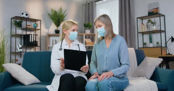 Healthcare έννοια όπου ελκυστική ειδικευμένη ξανθιά γυναίκα γιατρός σε μάσκα ακούγοντας για τα συμπτώματα της ασθένειας από τον ασθενή της κατά τη διάρκεια της επίσκεψης στο σπίτι της — Αρχείο Βίντεο