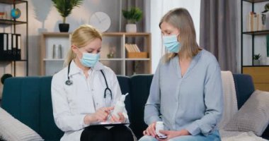Hasta kadın hastasına ev ziyareti sırasında nasıl hap ve yutucu kullanacağını açıklayan maskeli sarışın kadın doktorun ön görüntüsü.