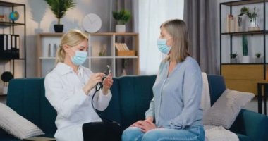 Çekici, kendine güvenen, yüksek yetenekli, koruyucu maskeli sarışın kadın-doktor. Ev ziyareti sırasında kalp ve ciğerleri kontrol eden 50 yaşında bir kadın.