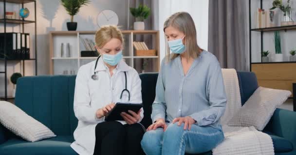 Atractiva y moderna doctora rubia experta en máscara y uniforme explicando los métodos de tratamiento utilizando información sobre tableta pc a su paciente femenina durante su visita en casa — Vídeo de stock