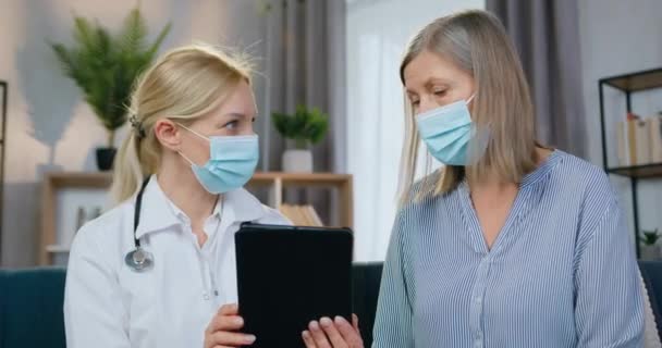 Портрет привабливої дружньої статі досвідченої жінки-лікарки в масці для обличчя, яка пояснює, як використовувати додаток для здоров'я для її пацієнта під час домашнього візиту — стокове відео