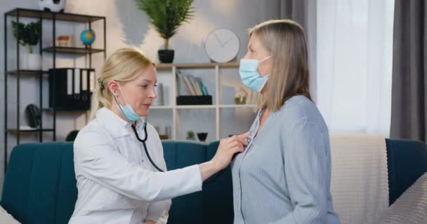 Довольно дружелюбная высококвалифицированная блондинка-врач проверяет сердцебиение у пациентки при помощи стетоскопа во время домашнего визита — стоковое видео