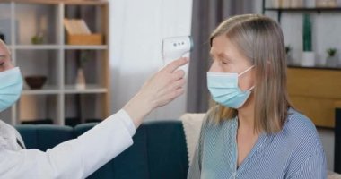 Sağlık hizmetleri ve tıp konsepti koruyucu maske takan çekici ve deneyimli kadın tıp çalışanı hastaların alnına temas edilmeyen kızılötesi termometre uygulayarak ısıyı kontrol ediyor.