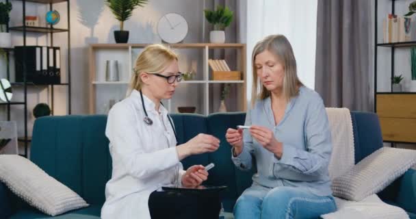 Belle amicale qualifiée blond femme-médecin donnant thermomètre à son patient pour mesurer la température lors de la visite à domicile, concept de soins de santé — Video