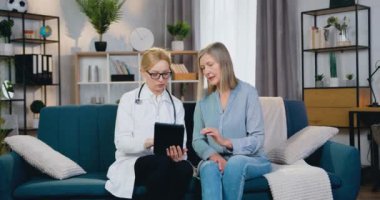 İyi görünümlü, kendine güvenen, deneyimli kadın doktor. Kadın hastasıyla koltukta oturuyor ve ev ziyareti sırasında sağlık uygulamasını tablet cihazda nasıl kullanacağını anlatıyor.