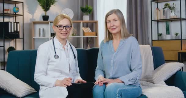 Портрет двох щасливих усміхнених дорослих жінок-лікарів і пацієнтів, які сидять на дивані і дивляться в камеру, коли лікар відвідує пацієнта вдома — стокове відео