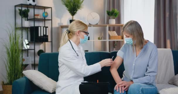 Pretty experienced female doctor in protective mask doing injection of vaccine to middle-aged woman in protective mask too when visiting her at home — Stock Video